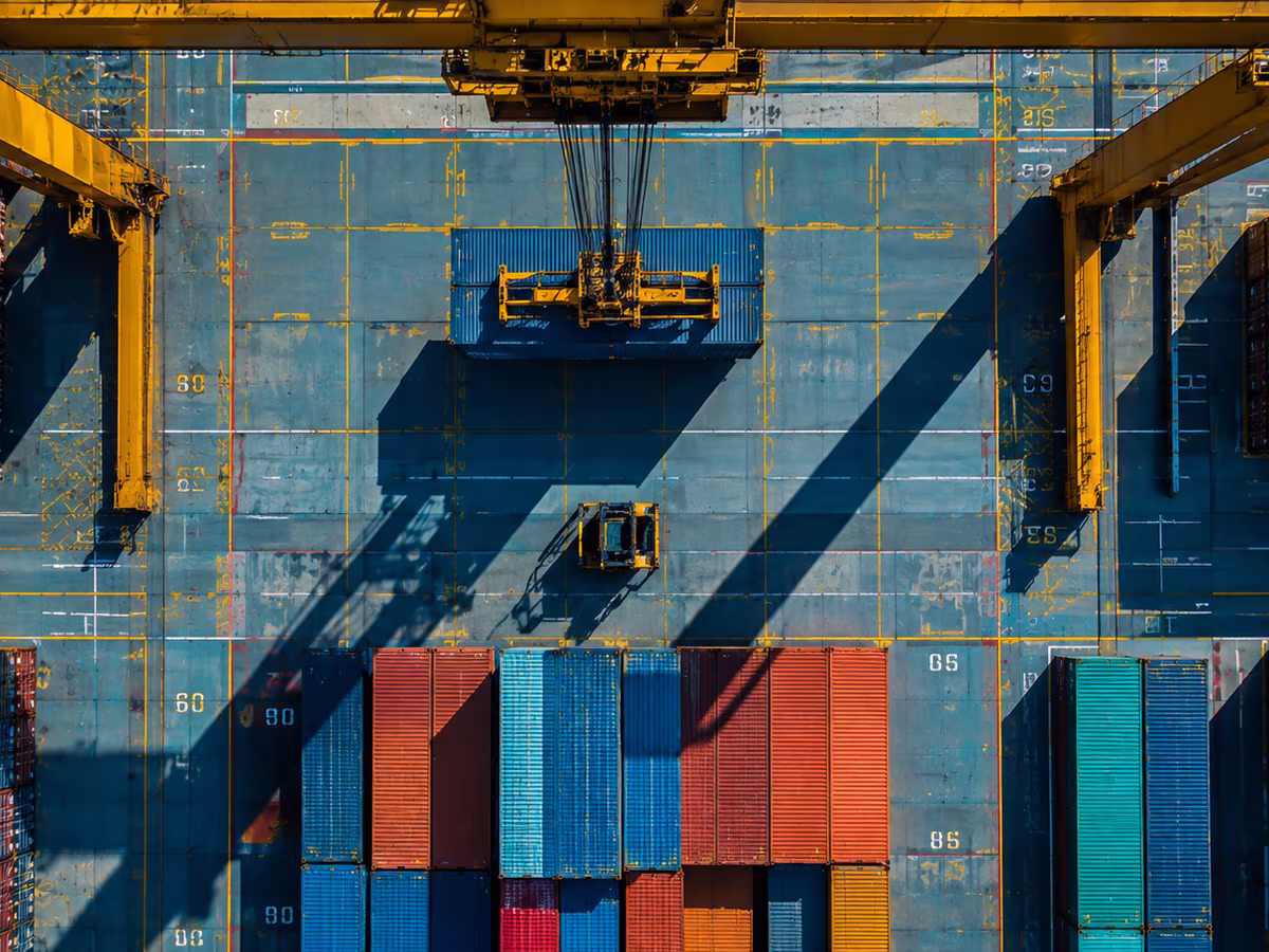 Stacked containers beneath a yellow gantry crane in the shipping yard